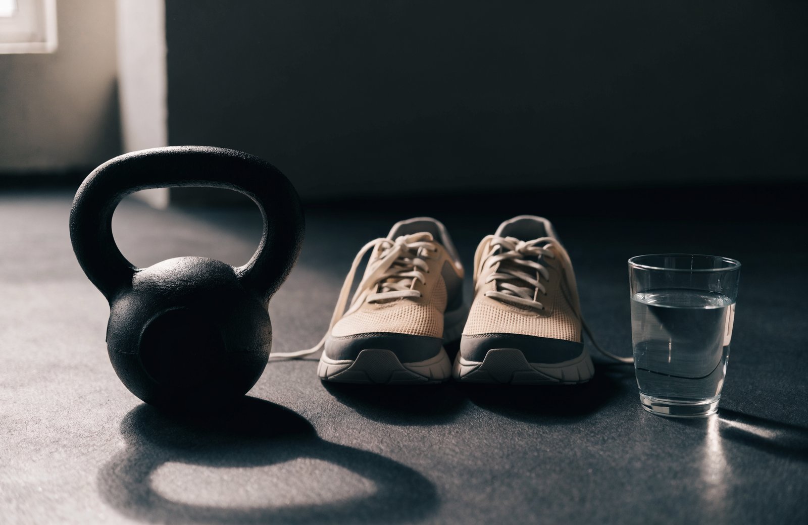 Kettlebell preto ao lado de um par de tênis de treino femininos e um copo de água no chão da academia